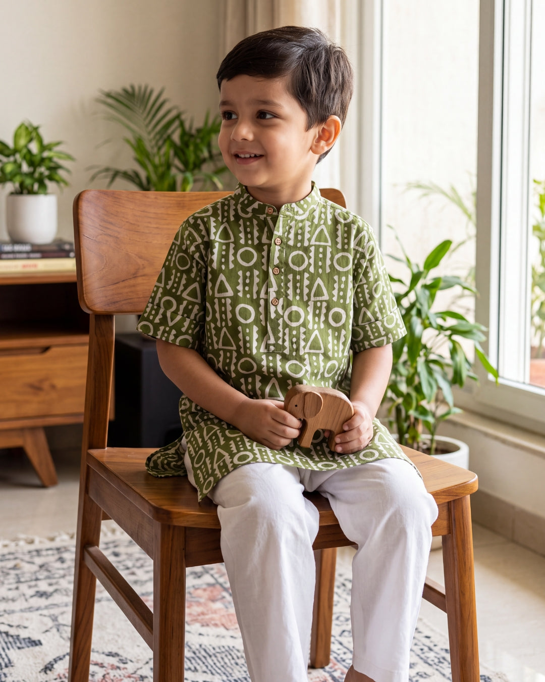 A young boy wearing अBIR Olive Green Virasat Long Kurta for Boys, white geometric patterned kurta and white trousers sits on a wooden chair in a bright room with plants and a window.