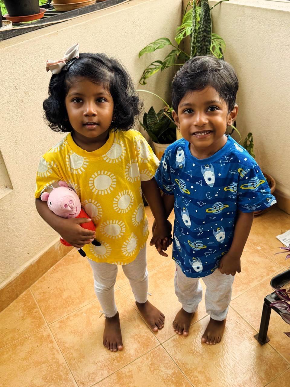 Two children holding hands on a tiled floor with plants in the background. From Sunshine Mornings to Space Adventures, 100% Organic Cotton All Over print kids tshirts from DoodleChic